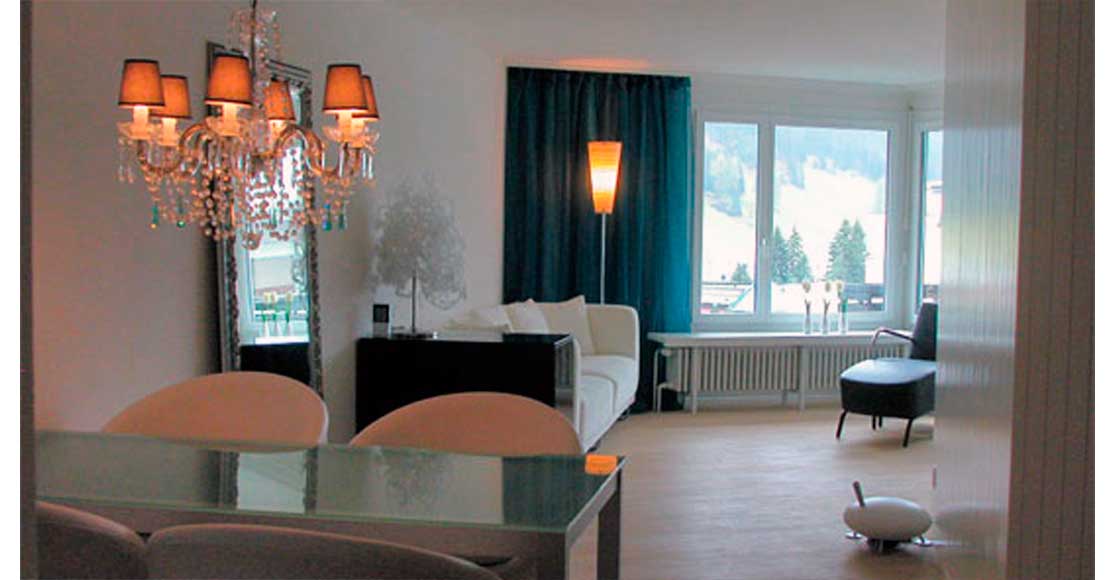 Foto: Wohn- und Essraum mit Tisch, weisen Stühlen, Kronleuchter, Spiegel, Lampen und Divan wie auch Sessel mit Hocker.